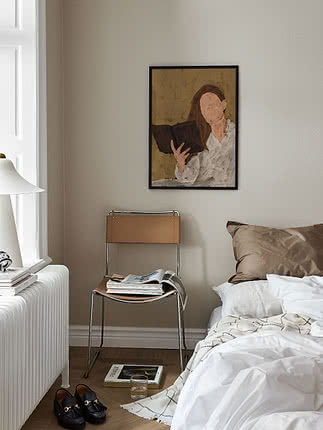 Dormitorio con cama, silla con revistas y cuadro enmarcado de una mujer leyendo en la pared