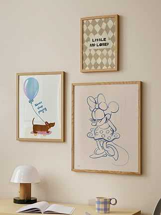 Tre indrammede børneplakater på beige væg over skrivebord med lampe, kop og åben bog.