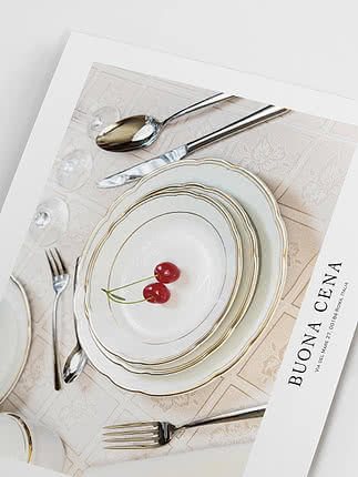 Elegante mise en place con piatti bianchi bordati d’oro, posate e due ciliegie.