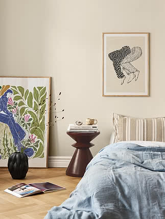 Hyggeligt soveværelse med lyseblåt sengetæppe, rundt sidebord, vase og indrammede kunsttryk.
