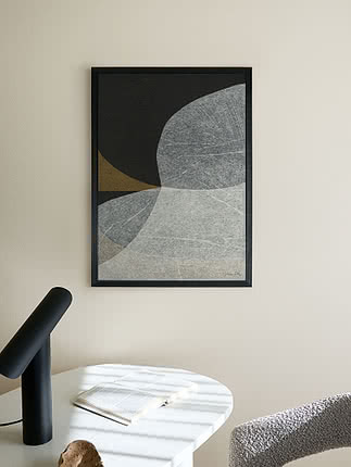 Abstrakt plakat i sort ramme over et hvidt skrivebord med sort lampe og en opslået bog