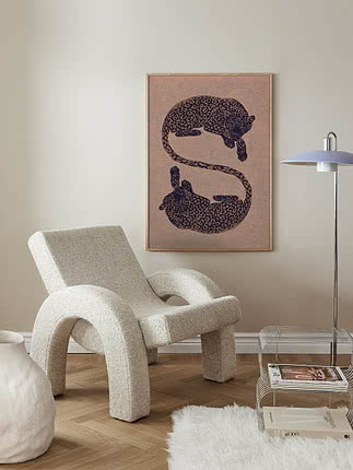 Postern Elegant Leopards i ekram på benvit vägg ovanför en vit boucléfåtölj, med keramikvas, fårskinnsmatta och kromat tidningsställ