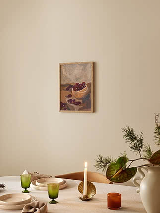 Red Cherries dans un cadre en chêne sur un mur beige au-dessus d'une table dressée : lin, grès crème, verres verts cannelés et bougeoir coquille en laiton