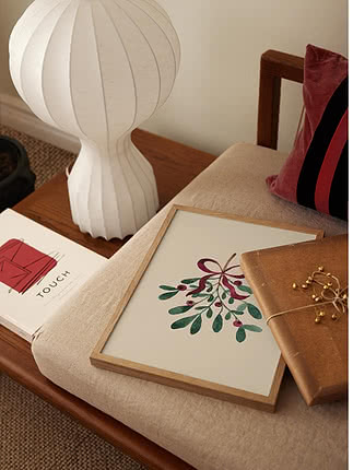 Festive Mistletoe dans un cadre en chêne posé sur un banc habillé de lin, près d'une lampe en papier cannelée, d'un paquet cadeau et d'un coussin en velours lie-de-vin