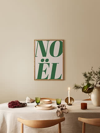 Noël dans un cadre en chêne sur un mur beige au-dessus d'une table dressée : lin, grès crème, verres verts cannelés, bougeoir en laiton et branches de pin