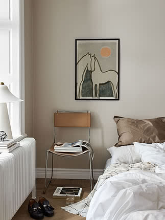 Plakaten Equine Embrace i sort ramme på beige soveværelsesvæg over en stol i cognacfarvet læder med magasiner, ved siden af en seng i hvidt lærred