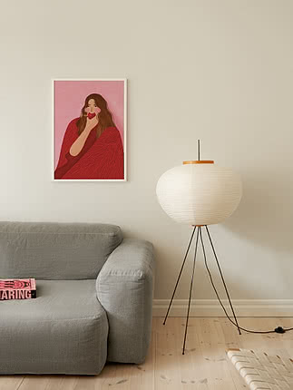 Plakaten Cozy Woman Apple i hvid ramme på en cremefarvet væg over en grå hørsofa ved siden af en gulvlampe i papir