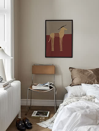 Poster Horse with Sombrero in cornice nera su parete beige di camera sopra una sedia in pelle carica di riviste, accanto a un letto rifatto in lino bianco