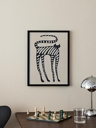 Póster Striped Whimsical Cat en marco negro en pared crema sobre un escritorio de madera oscura con un tablero de ajedrez a medio juego, un vaso de agua y una lámpara de mesa cromada