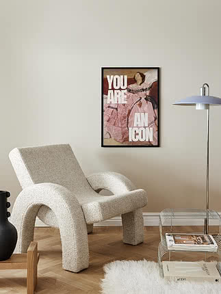 Poster You are an Icon in cornice nera su parete bianco sporco sopra una poltrona in bouclé crema, accanto a una piantana e un portariviste in filo