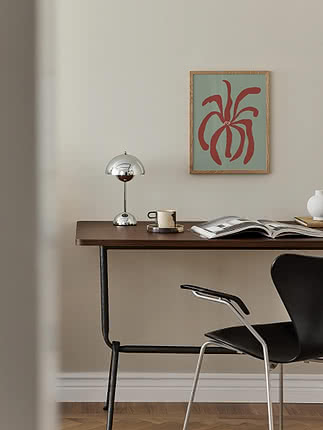 Muted Organic Bloom poster in a light oak frame on a pale wall above a dark wood desk with a chrome mushroom lamp, an espresso cup and an open magazine, beside a black chair