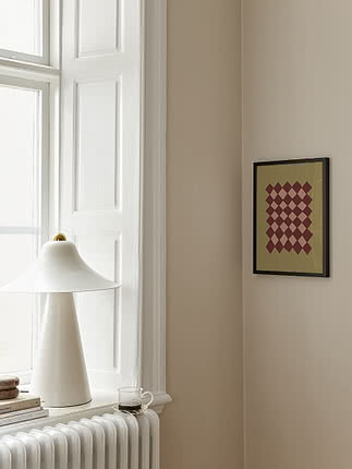 Póster Earthy Diamond Pattern en marco negro fino en pared beige en un rincón de ventana, junto a una lámpara blanca cónica en el alféizar y libros sobre un radiador