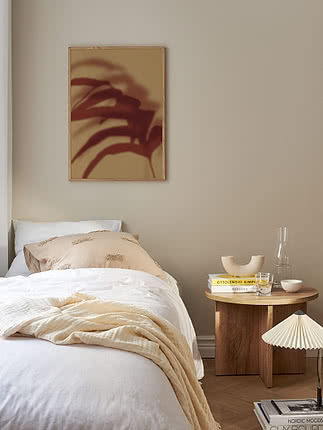 Plant Shadow poster in an oak frame on a greige bedroom wall above a bed with white and cream bedding, beside a round dark wood table with books, a pleated lamp and a glass