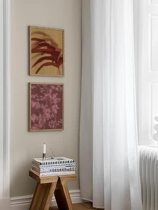 Botanical Shadows II poster in an oak frame hung below a second botanical print on a beige wall, beside a white curtain and a wooden stool with striped books and a candleholder