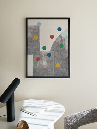 Plakat Geometric Play w czarnej ramie na kremowej ścianie nad białym okrągłym stołem z otwartą książką i drewnianym przedmiotem, obok czarna uchylna lampa i fotel z bouclé