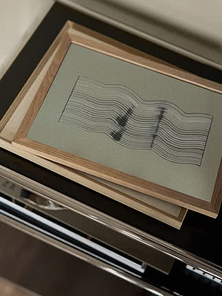 Poster Wavy Reflections I im Rahmen aus heller Eiche, auf einem Stapel Rahmen auf einer dunklen polierten Fläche liegend, schräg von oben gesehen