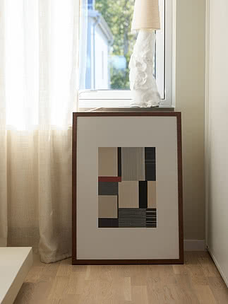 Plakat Geometric Harmony w ramie z ciemnego drewna oparty o ścianę pod oknem z lnianą zasłoną, obok biała fakturowana lampa na parapecie, na podłodze z jasnego drewna