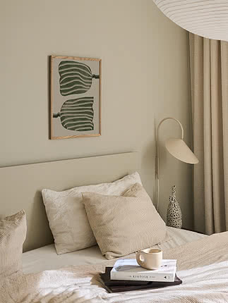 Abstract Tulips poster in an oak frame on a greige bedroom wall above a beige headboard with corduroy cushions, beside a cream wall lamp and a speckled vase on books