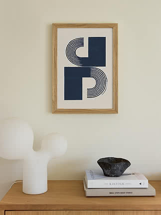 Plakat Modern Objects Blue w dębowej ramie na kremowej ścianie nad dębową komodą z białą rzeźbiarską lampą, stosem książek i misą z ciemnego kamienia