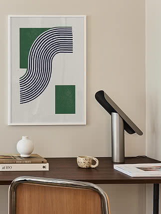 Plakat Modern Green Lines w białej ramie na kremowej ścianie nad orzechowym biurkiem z czarną uchylną lampą, okrągłym białym wazonem, książkami i cętkowanym kubkiem, obok krzesło z plecionką