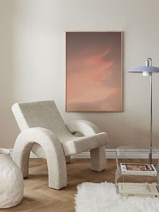 Plakaten Blush Sky i egeramme på en lys væg ved siden af en cremefarvet lænestol i bouclé, med en kromet gulvlampe, et magasinbord i metaltråd og et fåreskindstæppe på sildebensparket