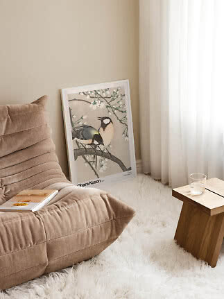 Inramad fågelposter lutad mot väggen bredvid beige fåtölj och ljust träbord.