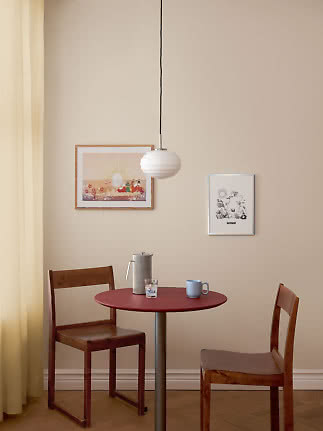 Spisehjørne med rundt rødt bord, to træstole og hvid pendellampe mod beige væg