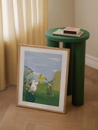 Framed Moomin poster leaning against the wall beside a round green side table