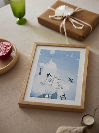 Affiche d’hiver Moomin encadrée sur une table avec cadeau, verre vert et demi-grenade.