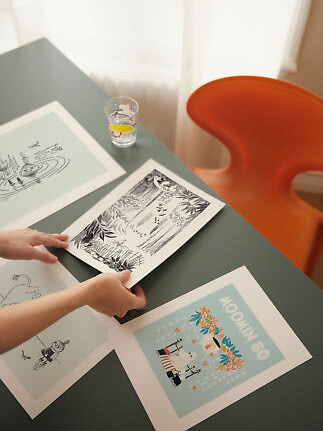 Hænder arrangerer illustrerede kunsttryk på et mørkt bord ved siden af et orange stol