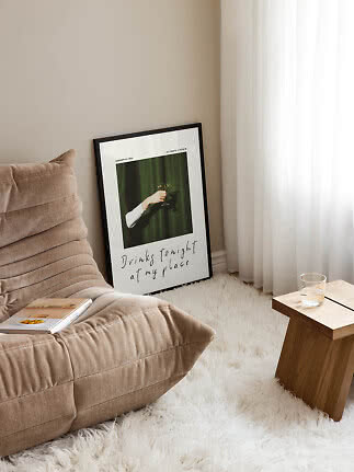 Indrammet plakat lænet mod væg ved beige loungestol, hvidt tæppe og lille træbord