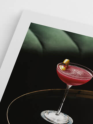 Rød cocktail i coupeglas med citronskal på sort bakke foran indrammet billede