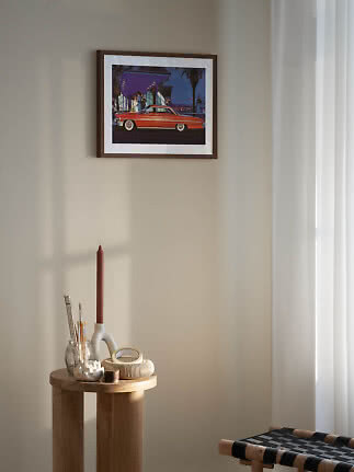 Framed retro car print on a light wall above a round wooden table with candle holder and decor