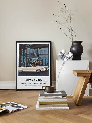 Framed vintage poster leaning against wall beside vase, books and wooden stool