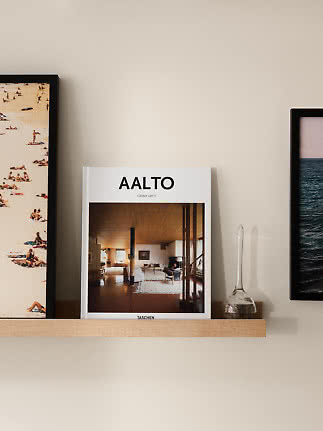 Album AALTO na drewnianej półce między oprawionymi grafikami i małym szklanym wazonem