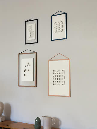 Four minimalist framed paper cut artworks hang on a light wall above a wooden table.