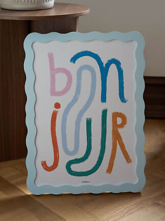 Colorful poster with abstract letter shapes in a wavy light blue frame, leaning against a wooden table