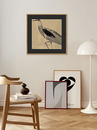 Poster con airone in passepartout nero e cornice in rovere su una parete beige, sopra uno sgabello di legno e una piantana