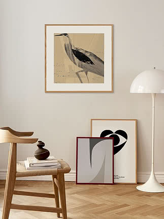 Poster con airone in passepartout bianco e cornice in rovere su una parete beige, sopra uno sgabello di legno e una piantana