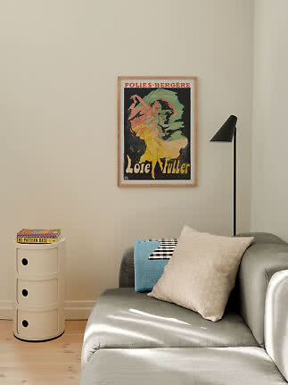 Indrammet Loïe Fuller-plakat over grå sofa med puder, sort gulvlampe og hvidt sidebord