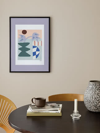 Framed abstract pastel print above a round table with a vase, candle holder, books and a mug.