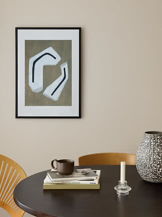 Framed abstract print with white shapes above a dining table with books, mug, candle and vase