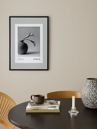 Black framed CALLA poster with black-and-white flower vase on beige wall above dark table