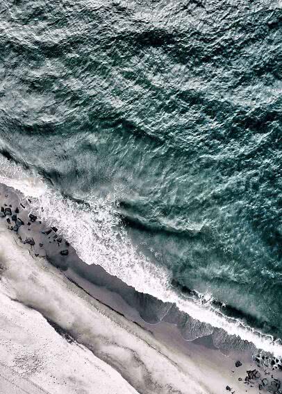 Aerial view of waves crashing onto a dark sandy beach with black rocks