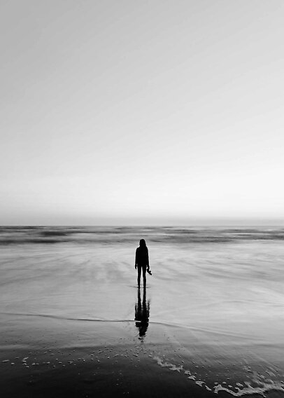 Silhuet af person på våd strand med sko i hånden, vendt mod havet i sort-hvid