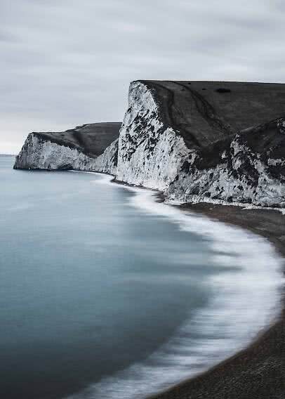 Acantilados blancos junto al mar con agua tranquila y playa curva bajo cielo gris