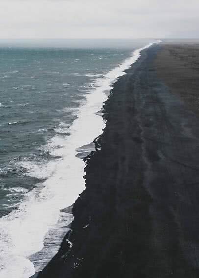 Luftfoto af bølger langs en lang sort sandstrand under overskyet himmel