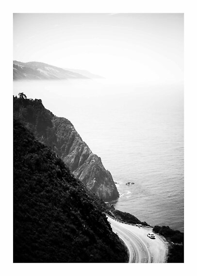 Black-and-white coastal landscape with winding road, car, cliffs, and calm sea in hazy light