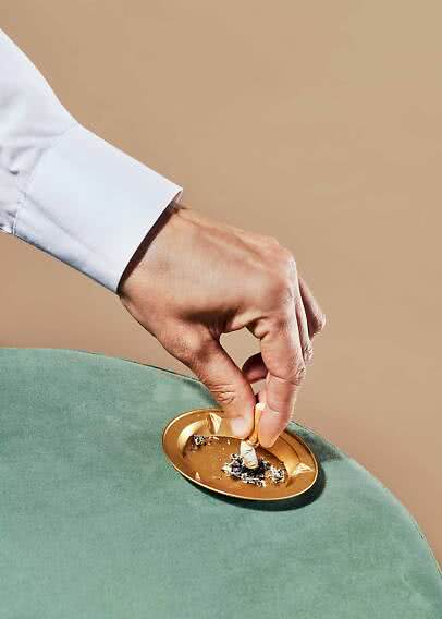 Mano con camicia bianca spegne una sigaretta in un posacenere dorato su sedia di velluto verde