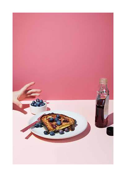 Toast topped with blueberries on a plate beside a bowl of blueberries and a bottle of syrup